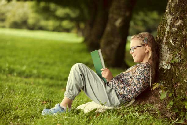 Fair-haired girl wearing spectacles reading her book under a tree.