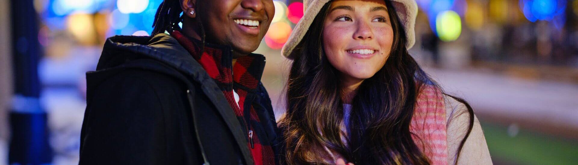 a smiling couple standing outside with blurred blue holiday lights in the backdrop, while the woman holds holiday shopping bags