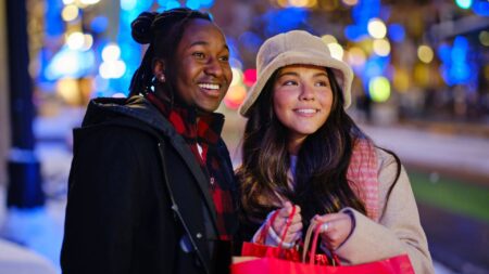 a smiling couple standing outside with blurred blue holiday lights in the backdrop, while the woman holds holiday shopping bags