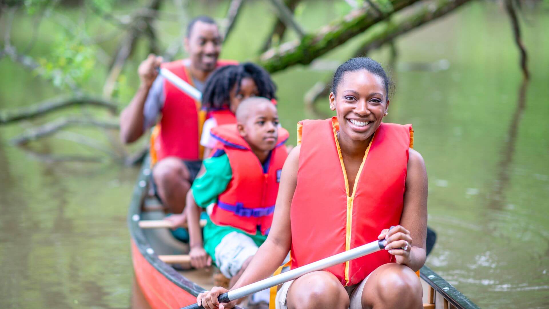 A family of four wearing life jackets while paddling a canoe in a river