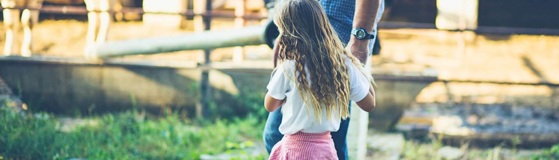 Young girl and adult explore a farm area with wooden fencing and outdoor equipment in warm autumn sunlight.